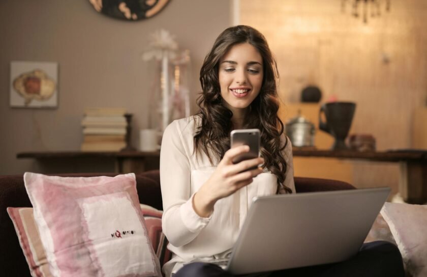 woman sitting on sofa while looking at phone with laptop on lap